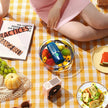 Person sitting on a yellow checkered blanket with a bowl of fruit, a book, and a small table with food.