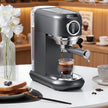 Espresso machine making a cup of coffee on a white surface with flowers and a glass of water in the background.