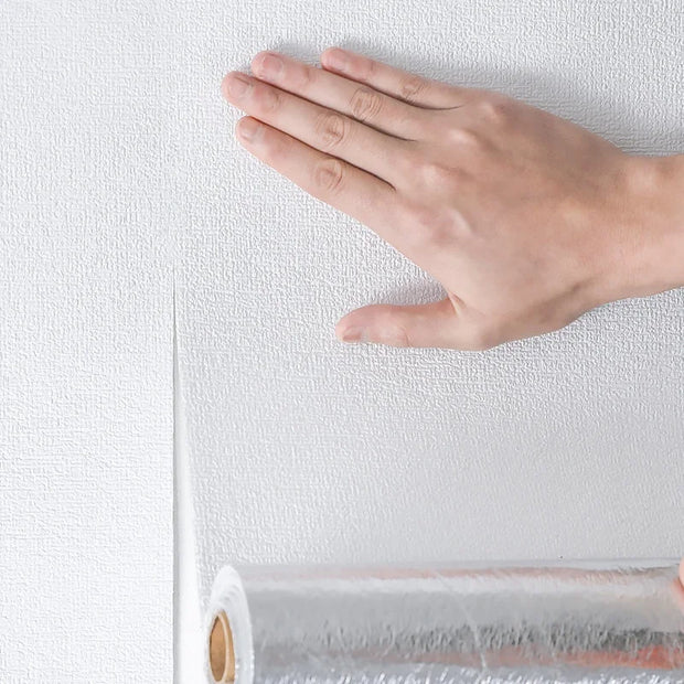 Hand touching a textured white wall next to a roll of wallpaper.