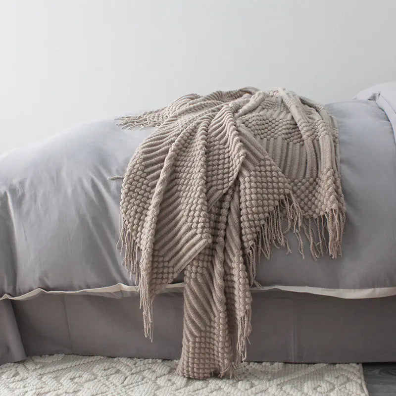 Beige knitted blanket with tassels draped over a gray bed.