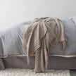 Beige knitted blanket with tassels draped over a gray bed.